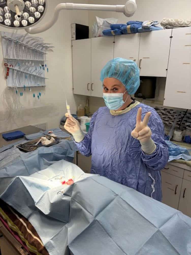 A surgeon in scrubs and a mask stands in an operating room, holding a syringe in one hand and making a peace sign with the other.