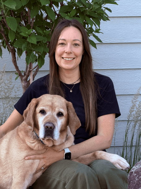 A woman with long brown hair sits outside in front of a light blue wall, smiling, with her arm around a large, light brown dog.
