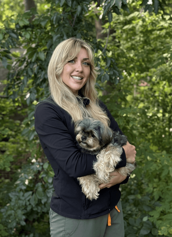 Woman with long blonde hair stands outdoors in front of green foliage, smiling and holding a small, fluffy black and gray dog in her arms.