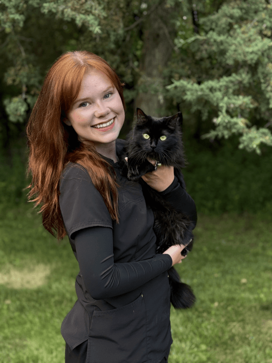 Woman with long red hair holds a fluffy black cat outdoors, standing in front of green trees and grass, both looking at the camera.
