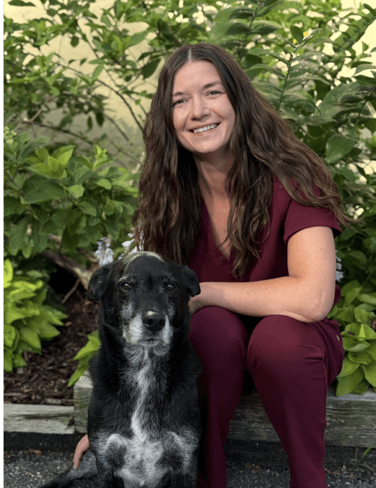 A woman in maroon scrubs sits outside on a bench next to a black and white dog, with green foliage in the background.