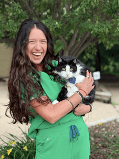 Woman in green scrubs stands outside, smiling and holding a black and white cat with a blue collar, near trees and plants.