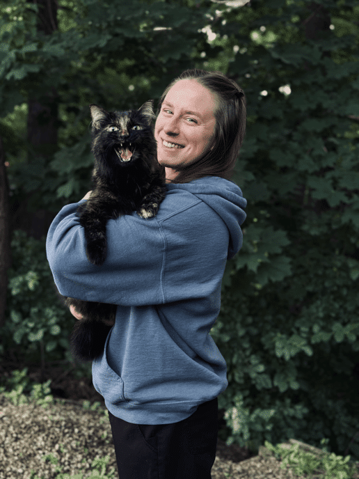 A person in a blue hoodie stands outdoors holding a black and brown cat, which appears to be meowing. Green foliage fills the background.