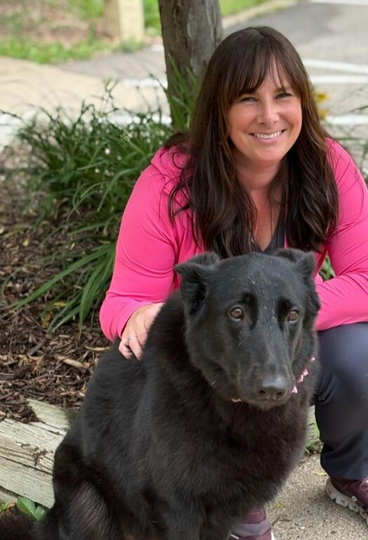 A woman in a pink jacket kneels beside a large black dog outdoors, both looking toward the camera.