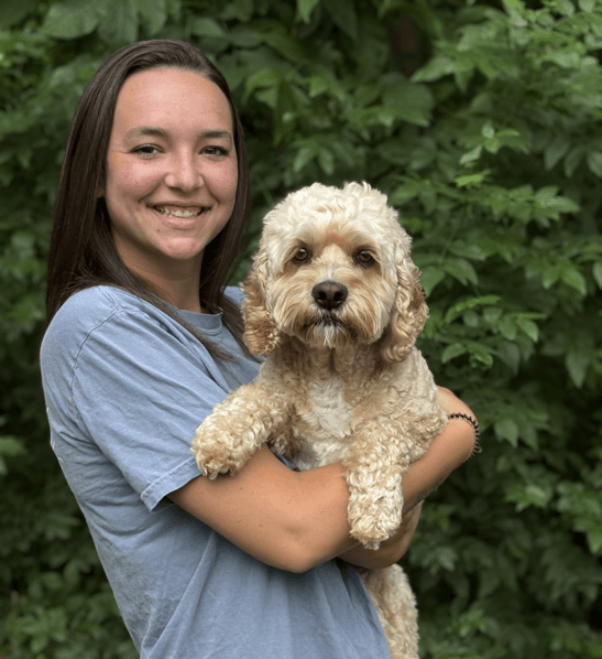 A person in a gray shirt smiles while holding a small, curly-haired dog in front of green foliage.